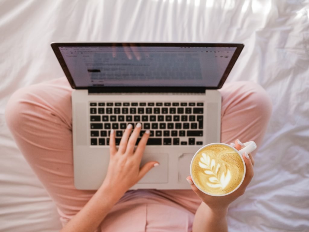 This is a picture of a woman using MacBook Pro and holding cappuccino. She is wearing a pink tracksuit and is sitting on a bed. The shot is from the top.